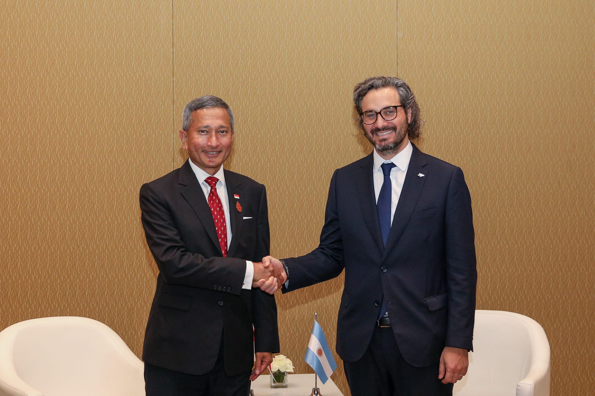 Two men in suits shake hands in front of a patterned wall, with an Argentinian flag visible.